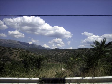 Yoğun yeşil ormanlarla kaplı dağlardan geçen yol mavi gökyüzünün altında bulutlarla kaplı..