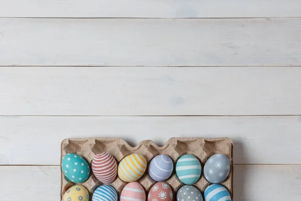 Una bandeja con coloridos huevos de Pascua hechos a mano en la superficie de madera blanca con espacio arriba. Estilo laico plano — Foto de Stock