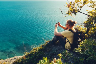 Gezgin adam kıyıda oturur ve cep telefonu kamera deniz günbatımı sırasında çekiyor