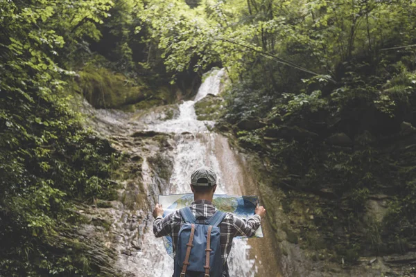 Explorador del viajero con la mochila de pie junto a la cascada y mira en el mapa Vista trasera vacaciones y turismo Concepto — Foto de Stock