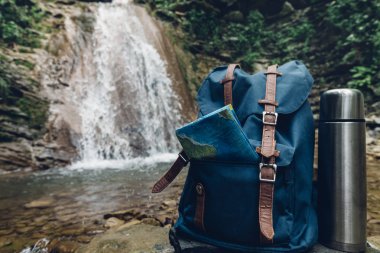 Hipster mavi sırt çantası, harita ve termos portre. Şelale arka plan üzerine açık turizm seyahat çantasından görünümü. Macera Hiking kavramı