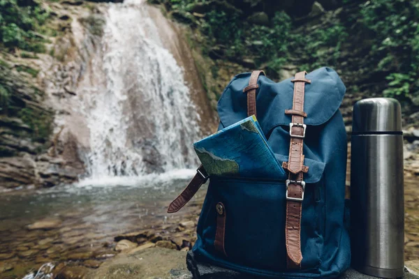 Hipster Blue Backpack, Map And Thermos Closeup. View From Front Tourist Traveler Bag On Waterfall Background. Adventure Hiking Concept