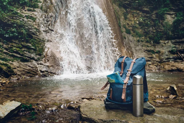 Hipster mochila azul, mapa y termos. Vista desde el bolso del turista delantero en fondo de la cascada. Wanderlast Adventure Concepto de senderismo — Foto de Stock