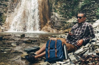 Backpacker adam içki çay veya kahve termosu kamp hedef seyahat kavramı dinlenme şelale arka plan üzerinde