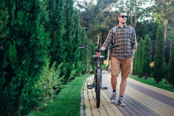 Hombre joven con gafas de sol caminando con bicicleta en el parque de verano — Foto de Stock