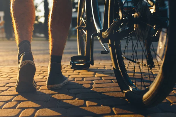 Las piernas de un hombre en zapatillas de deporte y una bicicleta de la ciudad se mueven alrededor de los guijarros contra el fondo de la luz del sol brillante durante la puesta del sol. Ángulo bajo, primer plano — Foto de Stock