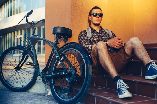 Guapo chico ciclista en gafas de sol con mochila sentado en los pasos cerca de la bicicleta y el descanso — Foto de Stock