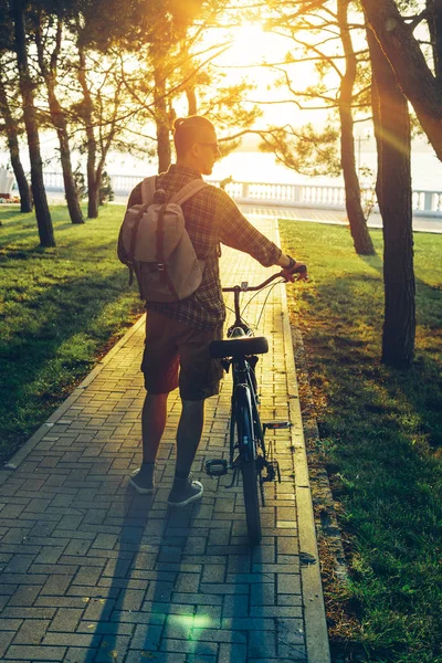 Hombre joven guapo caminando con bicicleta en el parque de verano al atardecer, actividad de fin de semana de vacaciones vista trasera — Foto de Stock