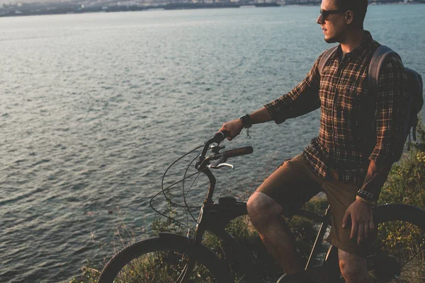 Joven hombre de pie con bicicleta en la costa y disfrutando de la vista de la naturaleza Puesta de sol vacaciones viajando Relajación Descanso Concepto — Foto de Stock