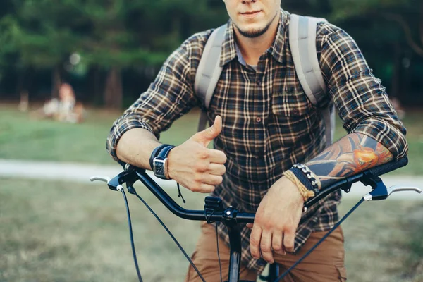 Hombre joven está haciendo pulgar hacia arriba por las manos sentadas en una bicicleta en el verde prado de verano — Foto de Stock