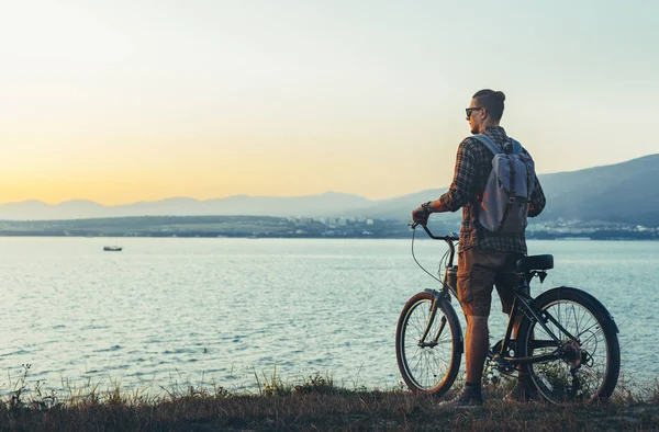 Joven ciclista de pie con bicicleta en la costa y disfrutando de la vista de las vacaciones al atardecer viajando Destino Descanso Concepto — Foto de Stock