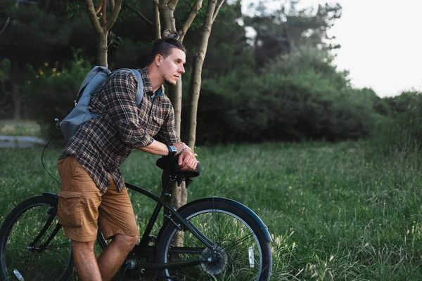 Guapo joven ciclista de pie descansando con bicicleta cerca del árbol en el parque de verano y descanso recreación viaje destino concepto — Foto de Stock
