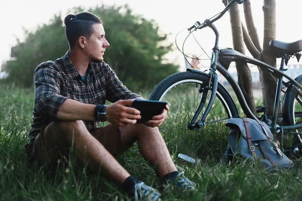 Joven ciclista viajero se sienta en el prado de verano cerca de la bicicleta, celebrando en la Tablet Recreación Descanso Destino de viaje Concepto — Foto de Stock