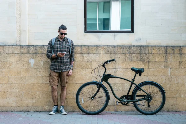 Guapo joven ciclista de pie junto a la bicicleta y su mirada en el teléfono inteligente. Street Lifestyle Concepto urbano cotidiano — Foto de Stock