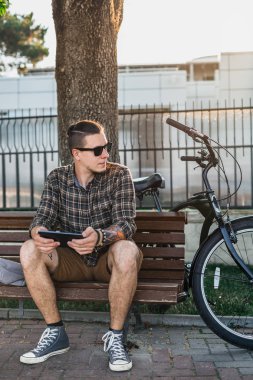 Yaz parkta bankta oturan ve Tablet iletişim bağlantı dijital cihazlar teknolojisi kavramı kullanarak bisikletçi genç adam