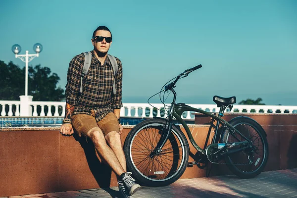 Ung Man i solglasögon sitter på fontänen bredvid cykel i sommar Park dagliga livsstil Urban vilar koncept — Stockfoto