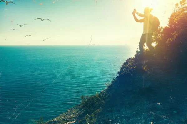 Мандрівник стоїть зверху і фотографує море на камеру мобільного телефону під час заходу сонця вінтажне тоноване зображення — стокове фото