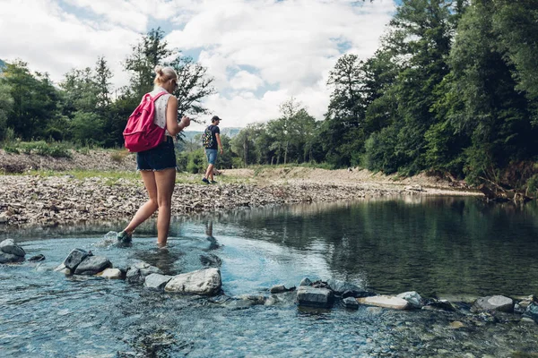 Pareja joven caminando a lo largo de la costa del río de montaña en verano. Chica vadeando a través del río — Foto de Stock