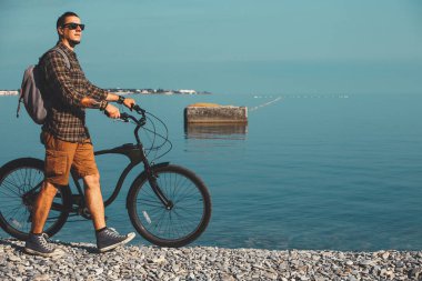 Genç bisikletçi kıyısında yürüyüş ve deniz görünümü zevk bisiklet ile güneş gözlüklü adam. Tatil seyahat etkinlik kavramı