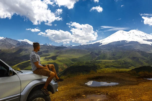 El joven viajero se sienta en el coche y disfruta de la vista de las montañas en verano. Región de Elbrus, Cáucaso Septentrional, Rusia — Foto de Stock