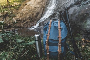 Hipster mavi sırt çantası, termos ve Trekking Polonyalılar portre. Şelale arka plan üzerine açık turizm seyahat çantasından görünümü. Macera Hiking kavramı