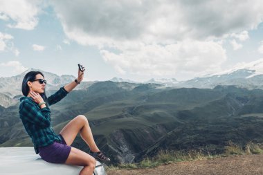 Genç kız araba ve yapar Selfie üzerinde Smatrphone yaz dağ zemin üzerine oturur. Seyahat keşif yolculuğu kavramı