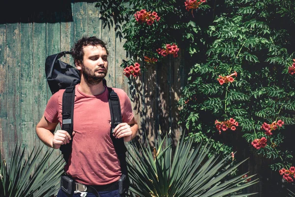 Joven viajero hombre con mochila de pie cerca de la vieja pared de madera con plantas — Foto de Stock