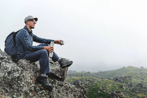 Hombre viajero joven sentado en la parte superior y sosteniendo termos en su mano. Senderismo Aventura Concepto de Turismo — Foto de Stock