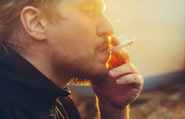 Porträtt av stilig ung Man med skägg som röker cigarett mot bakgrund av Bright Sun, närbild — Stockfoto