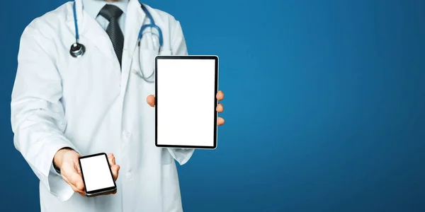 Doctor sostiene Smartphone y tableta digital con espacio de copia y ruta de recorte para la pantalla en fondo azul —  Fotos de Stock