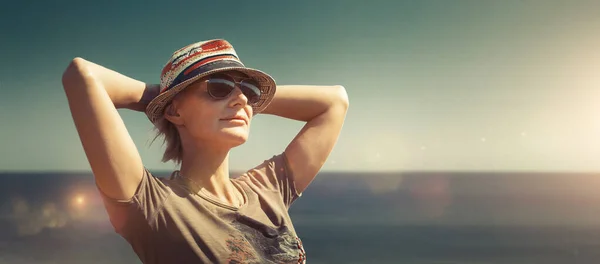 Özgürlük, mutluluk, turizm, macera ve boş zaman kavramı. Çekici orta yaşlı şapkalı kadın, ellerini başının üstüne koy ve güneşli ışıkta tatil köyünde manzaranın tadını çıkar.