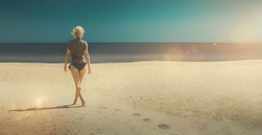 Güzel, zayıf, sarışın, orta yaşlı kadın sahilde yürüyor ve okyanusa bakıyor. Güneşli bir ışıkla dikiz manzarası. Özgürlük, seyahat, macera, temiz gökyüzü geçmişi, doğal minimalizm kavramı. Metin için resim pankartı