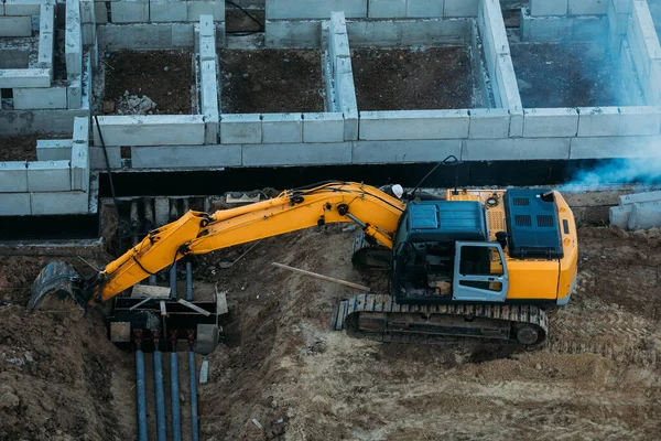 住宅の基礎の隣の建設現場で働く黄色い掘削機のトップビュー — ストック写真