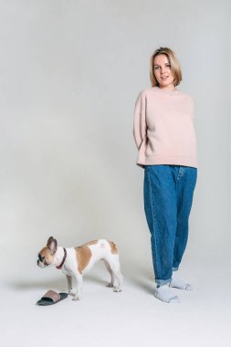 Pembe stüdyo süveterli, beyaz arka planlı genç kız komik Fransız buldoguyla oynuyor.