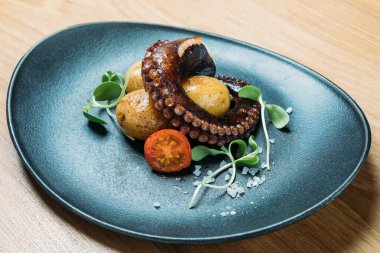 Izgara ahtapot dokunaçları, patates ve sebzeyle servis edilmiş domatesler bir restoranda mavi bir tabakta.