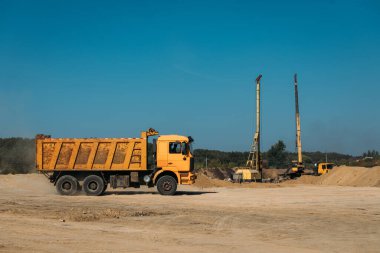 Sarı damperli kamyon, güneşli bir günde kumsalda bir inşaat alanında arka planda bir kazık sürücüsüne karşı.