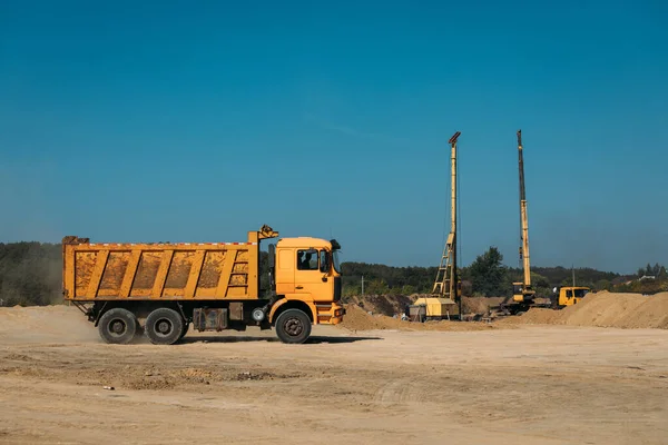 Sarı damperli kamyon, güneşli bir günde kumsalda bir inşaat alanında arka planda bir kazık sürücüsüne karşı.