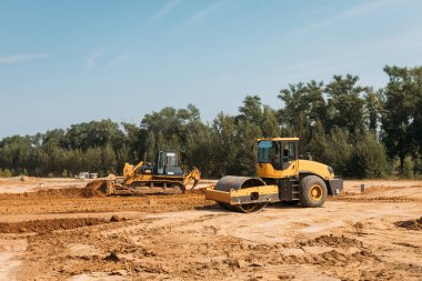 Sarı asfalt kaldırıcı sıkıştırıcısı güneşli bir günde bir inşaat alanında kumlar üzerinde çalışıyor.