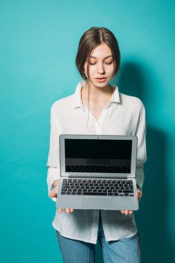 Stüdyoda mavi arka planda dizüstü bilgisayarı olan beyaz tişörtlü ve kot pantolonlu genç bir kız.