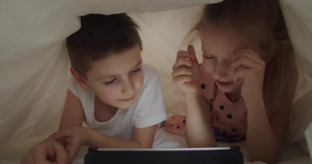 Niño y niña acostados debajo de la manta con la tableta — Vídeo de stock