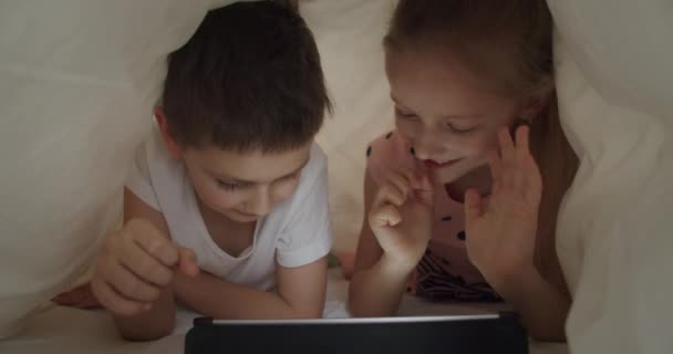 Niño y niña acostados debajo de la manta con la tableta — Vídeo de stock