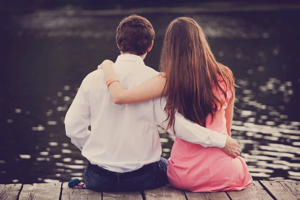 Romantisk sommar dag utomhus. Ungt par i kärlek, sitter embracing, på en träbrygga vid sjön och ser på vattnet. Bakifrån — Stockfoto