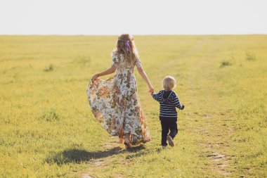 Genç anne ve küçük oğlu elele alanında üzerinde yürüyen güzel görünümünü geri yaz gün. Mutlu aile kavramı
