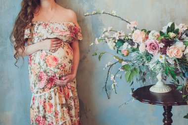 Güzel renkli giysili bir hamile kadına iç karın evde çiçek ve Beklemeler ellerinde parlak bir buket yanında duruyor. Gebelik, ebeveynlik, hazırlık ve beklenti kavramı