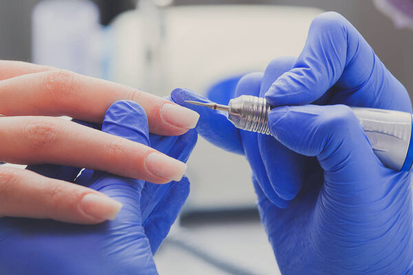 Closeup shot of manicurist in blue rubber gloves cleans cuticle on female nails using a milling cutter for manicure. Professional manicure and nail care procedure in beauty salon