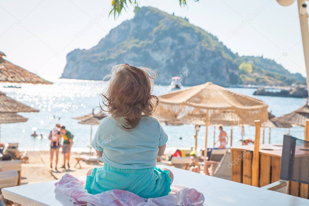 Amazing view of little girl sitting on a table and looking on turquoise seaside in sunny summer day, Corfu, Greece