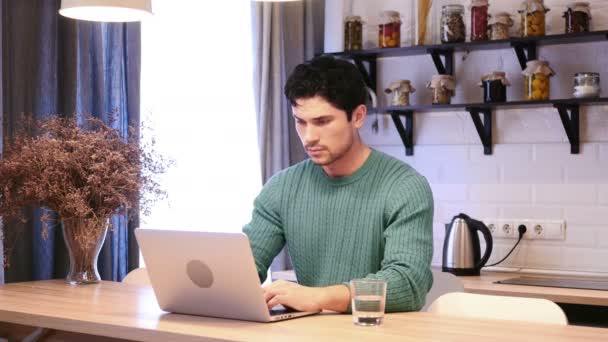 Молодой человек с болями в спине работает над ноутбуком на кухне — стоковое видео