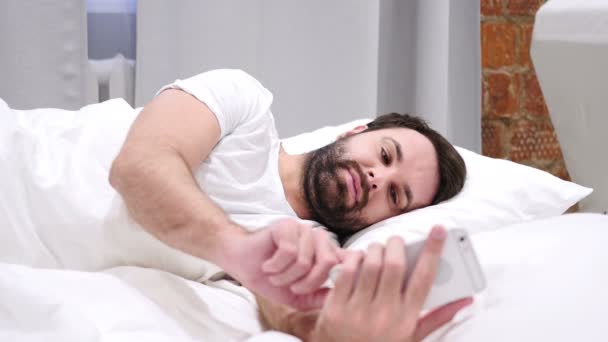 Barba hombre acostado en la cama en el lado de la navegación en el teléfono inteligente por la noche — Vídeos de Stock