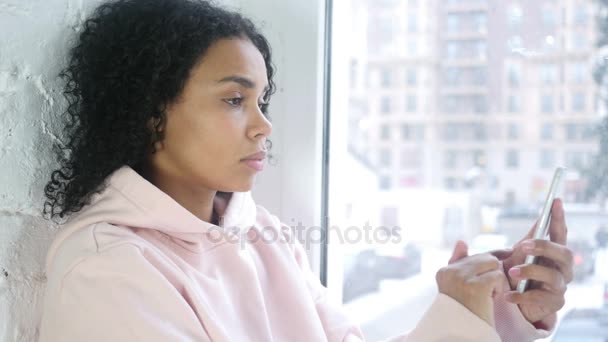 Mujer Afroamericana Navegando Smartphone, Sentada en la Ventana — Vídeo de stock