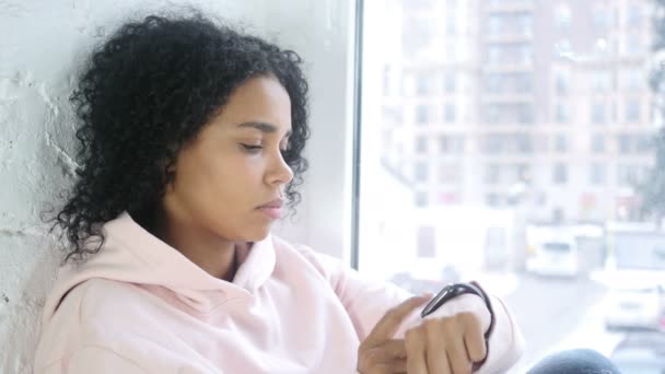 Afro-American kvinna använder Smartwatch för bläddring, sitter vid fönstret — Stockvideo